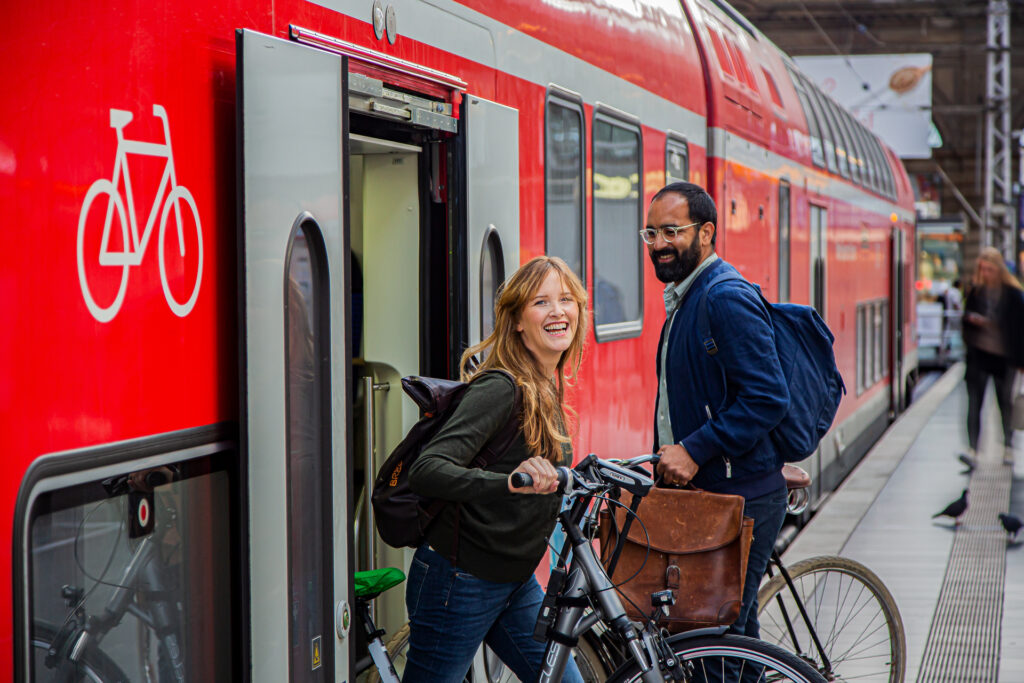 Eine Frau und ein Mann steigen mit ihren Fahrrädern in den Zug ein bzw. aus dem Zug aus.