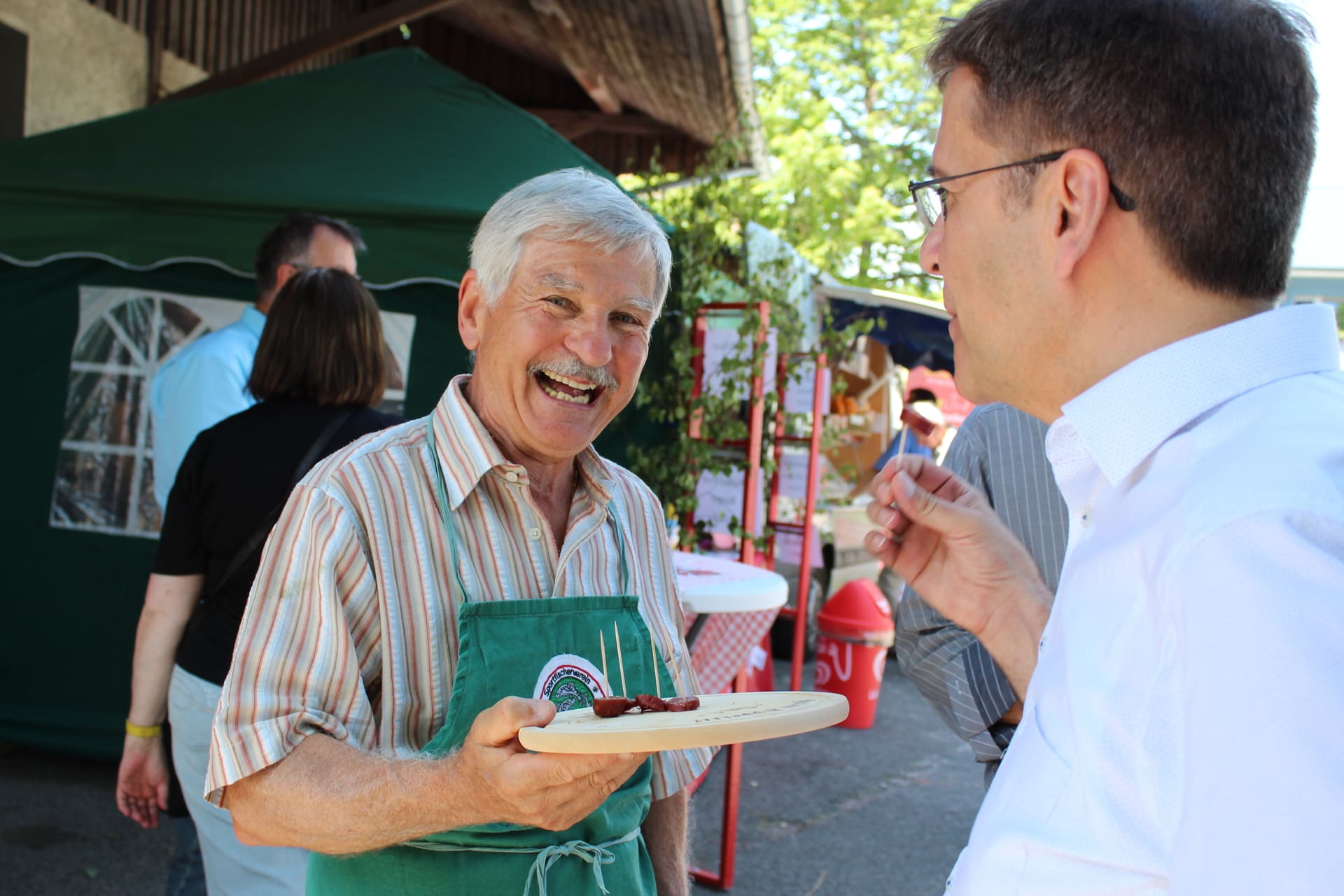 Lachender Mann hält Brettchen mit kleinen Wurststückchen zum Probieren bereit